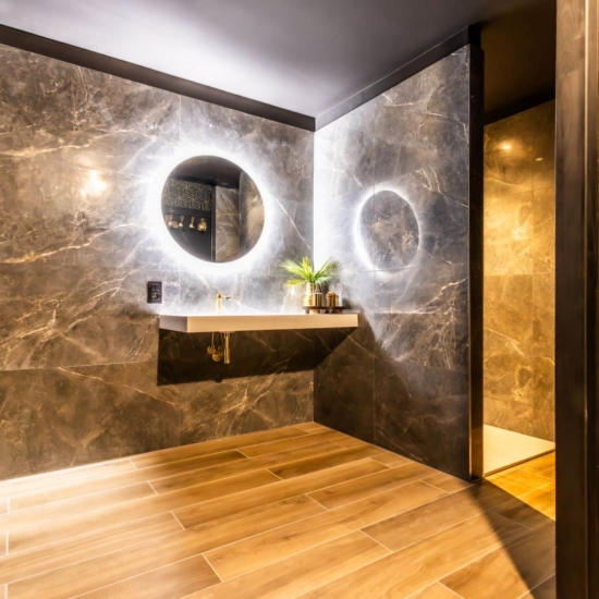 Salle de bain avec revêtement en marbre, miroir circulaire rétroéclairé, et lavabo suspendu dans une suite privative